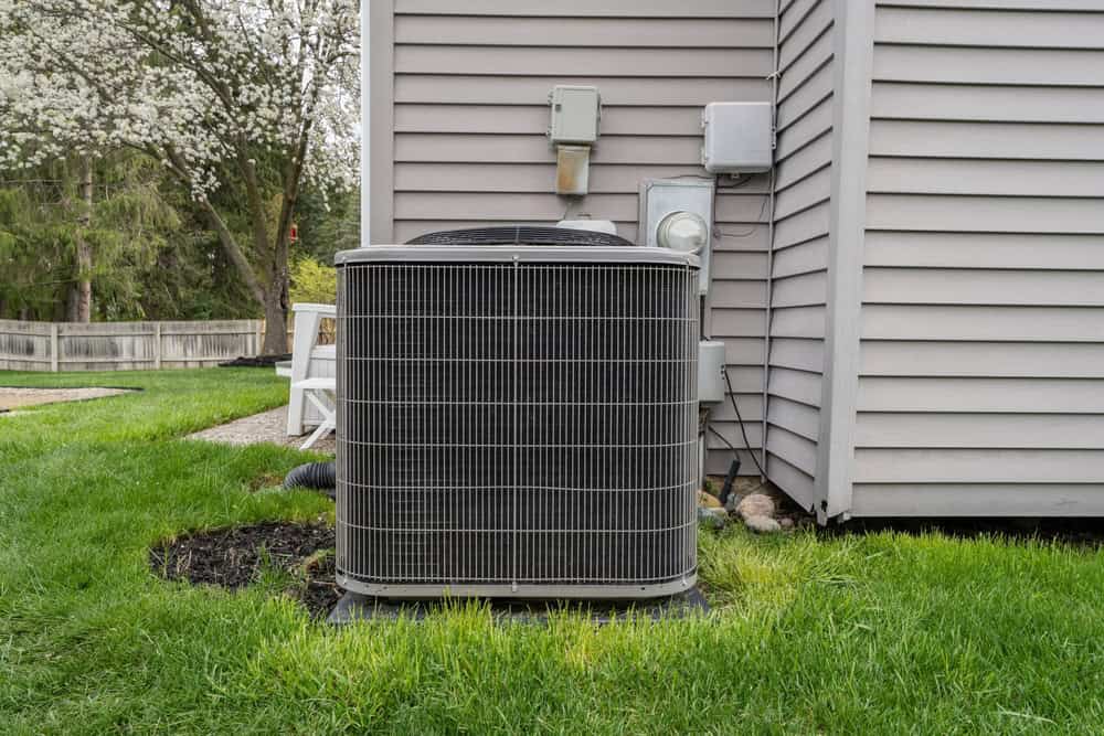 An air conditioning unit is positioned on a concrete slab next to the exterior wall of a house, surrounded by green grass. A tree with white blossoms is visible in the background.