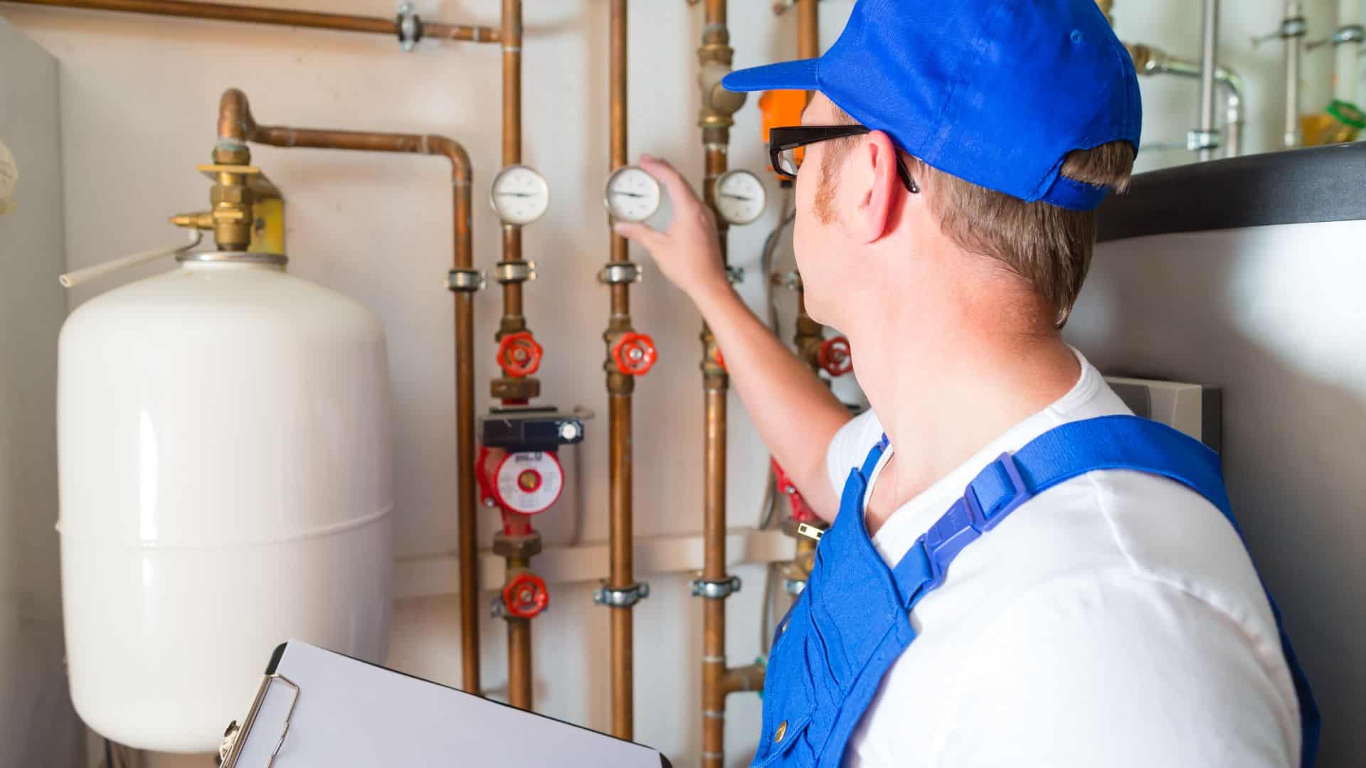 A plumber in a blue uniform and cap checks pressure gauges and pipes in a utility room. He holds a clipboard while adjusting a valve on the pipe system, which includes a white tank and several red valves.
