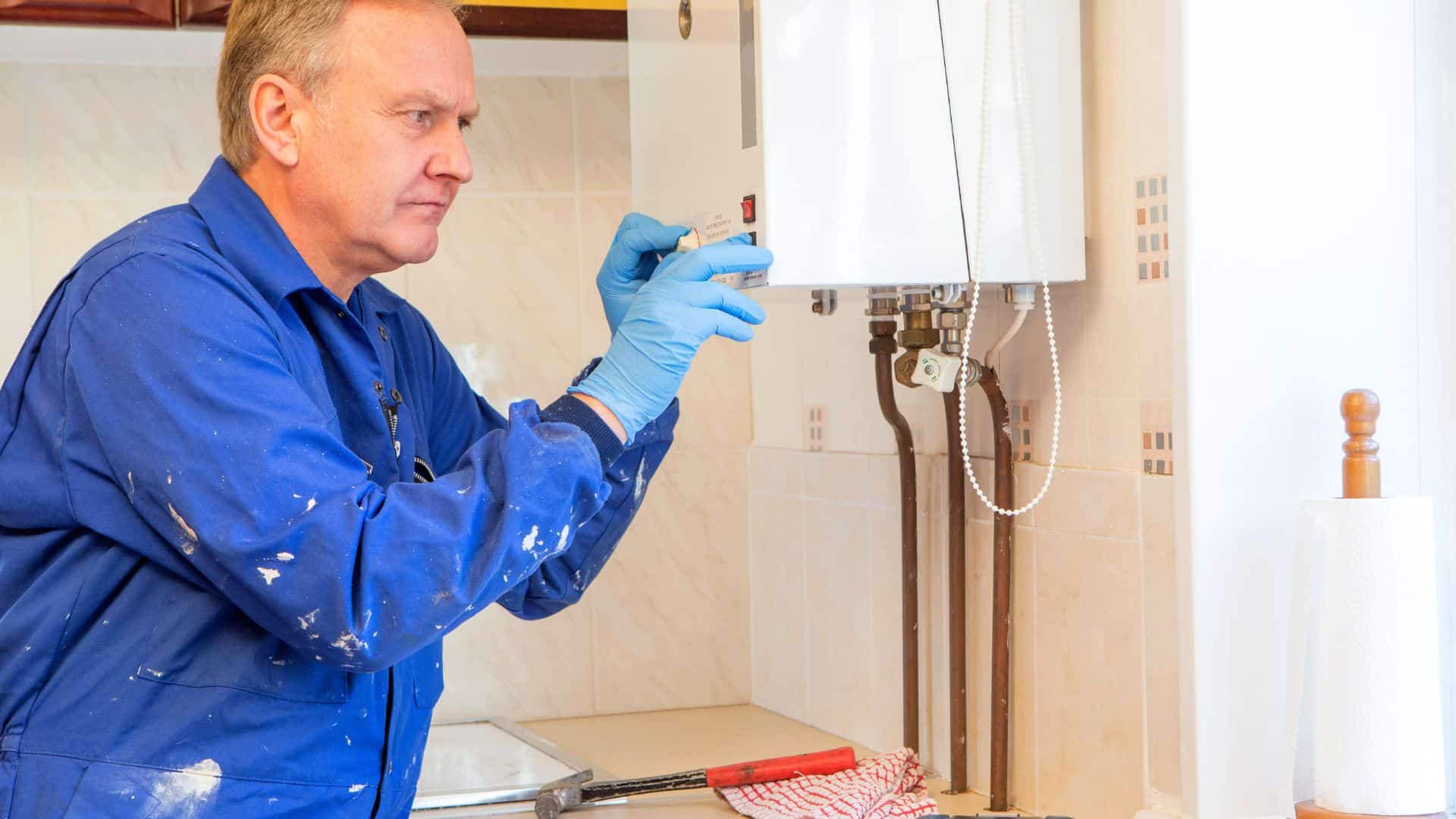 A man in a blue jumpsuit and gloves is inspecting a wall-mounted boiler in a well-lit kitchen. He is carefully adjusting a component, with tools and a red cloth on the countertop nearby.