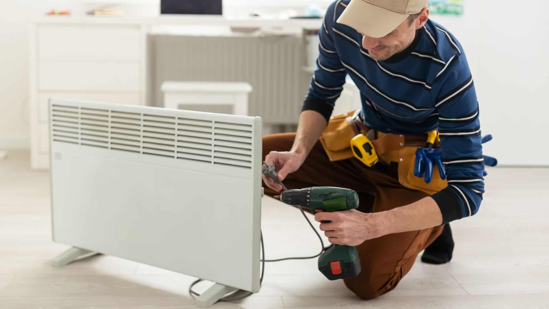 A person wearing a cap and striped sweater uses a power drill to work on an electric heater. They are kneeling on the floor in a bright room and have tools in a belt. The setting appears to be a home or office.