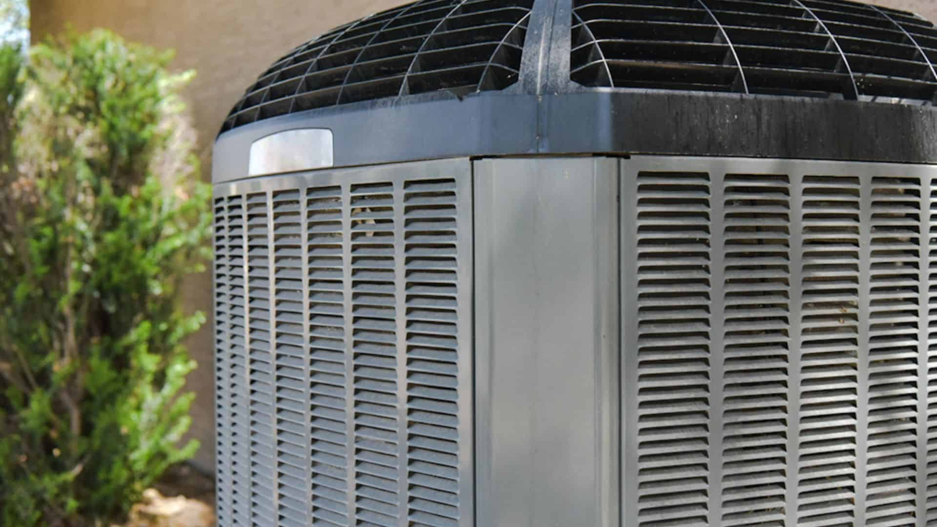 Close-up of an outdoor air conditioning unit with a grille and fan on top, situated near shrubbery. The unit has a metallic finish, and the surroundings suggest it is installed in a residential setting.