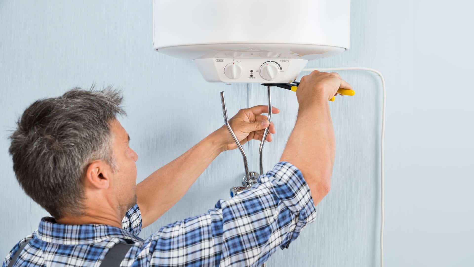 A man in a plaid shirt uses a wrench to adjust a white water heater attached to a light blue wall.