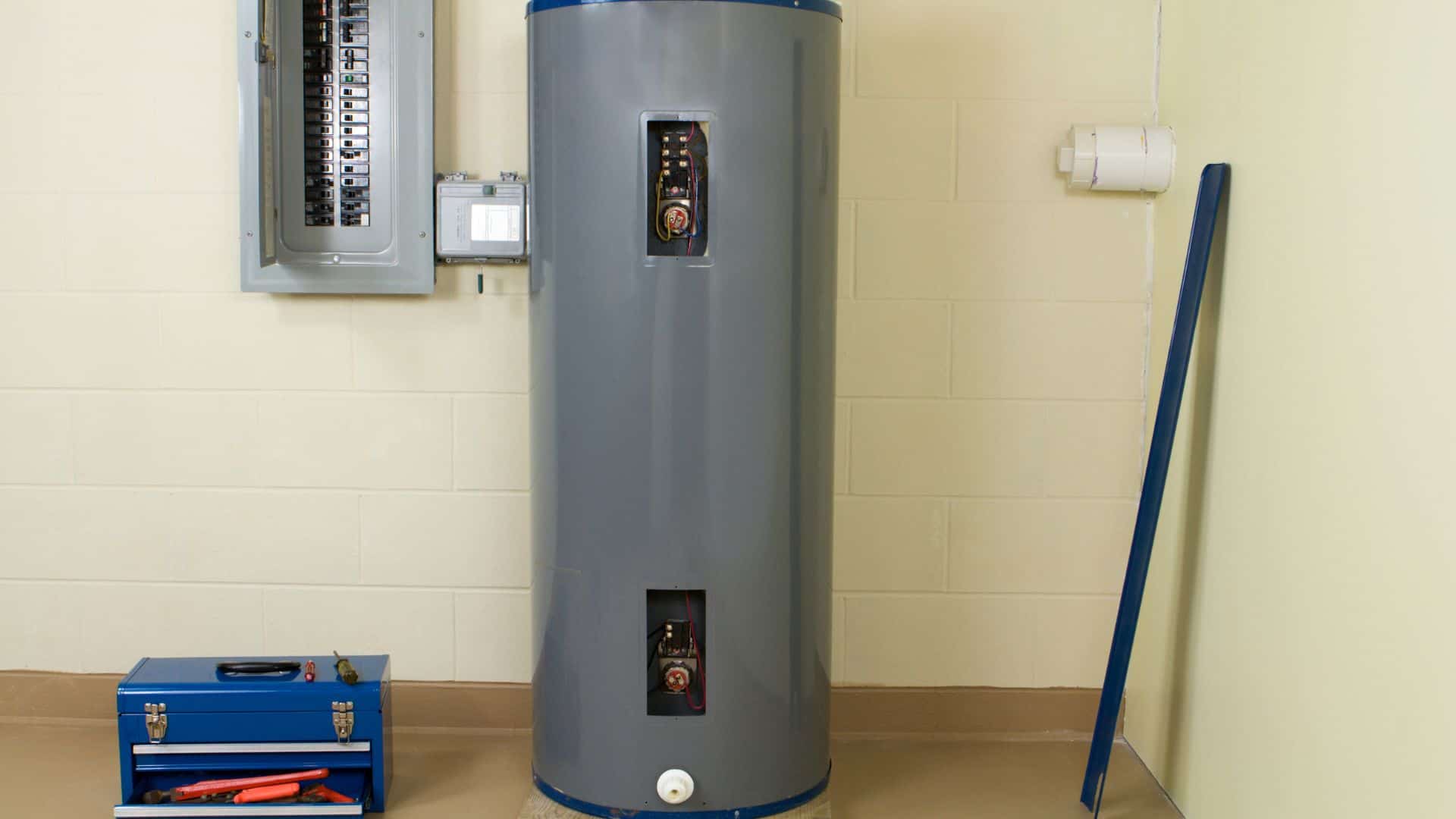 A gray water heater stands in a utility room with light beige walls. An electrical panel and a blue toolbox with tools are nearby. A long blue object leans against the wall.