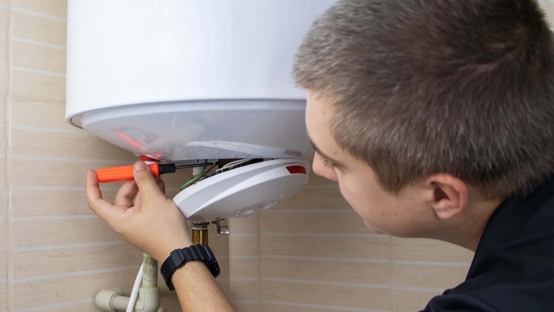A person with short hair wearing a black shirt is using a screwdriver to repair or install a white water heater mounted on a tiled wall. The person is focusing on fixing the panel beneath the heater.
