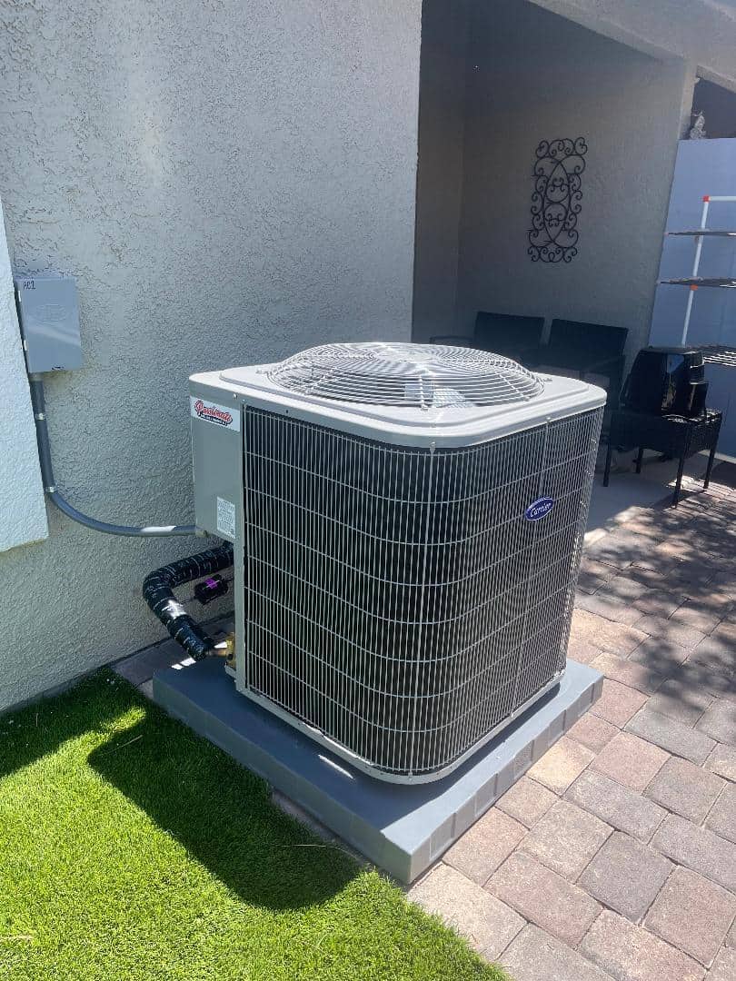 A large outdoor air conditioning unit sits on a concrete pad next to a white building. The area is landscaped with green grass and a paved walkway. There are chairs and a decorative wall piece visible in the background.