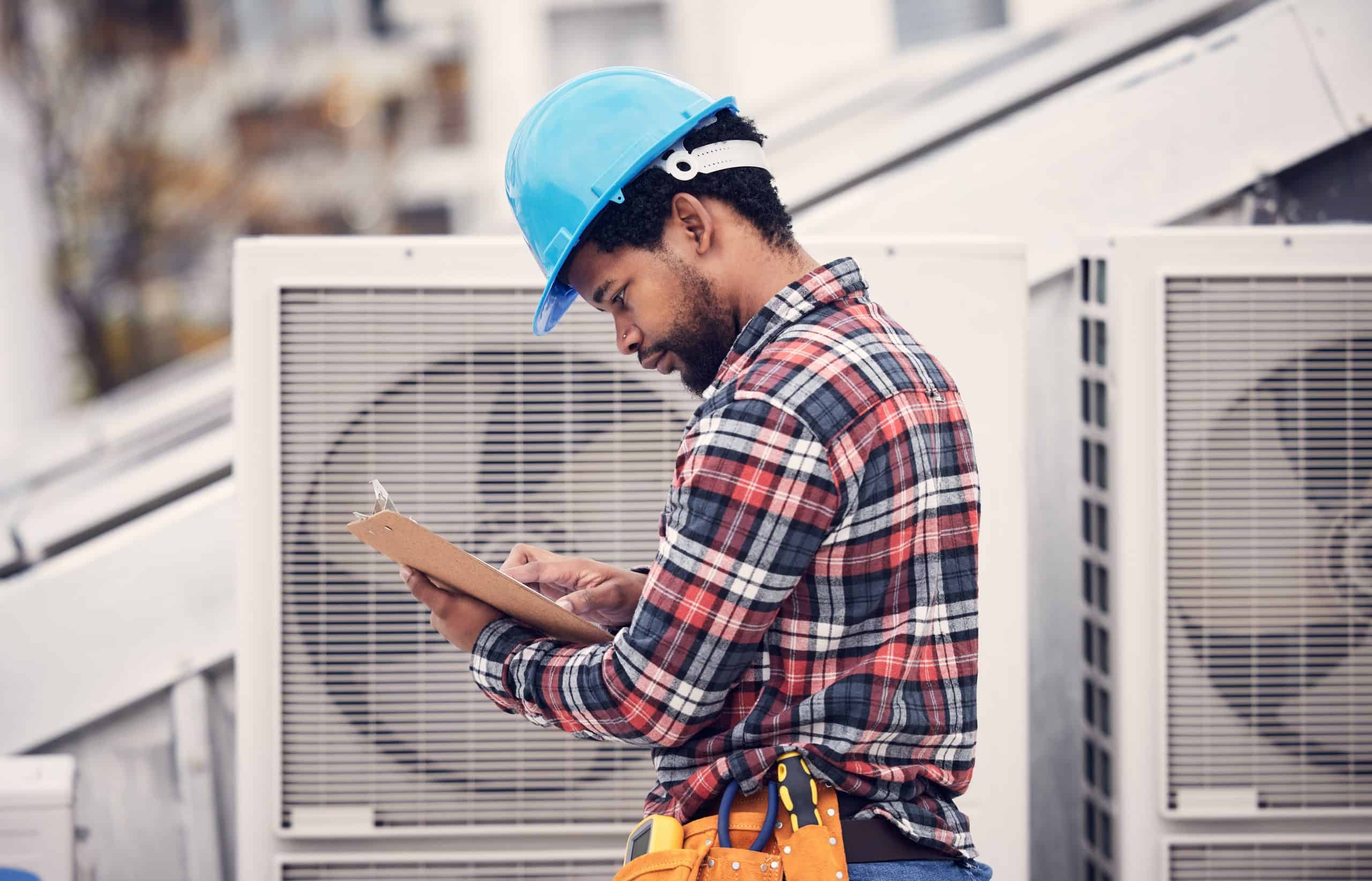 A construction worker in a blue hard hat and plaid shirt checks a clipboard while standing next to industrial HVAC units on a rooftop. He wears a tool belt and focuses on his task.