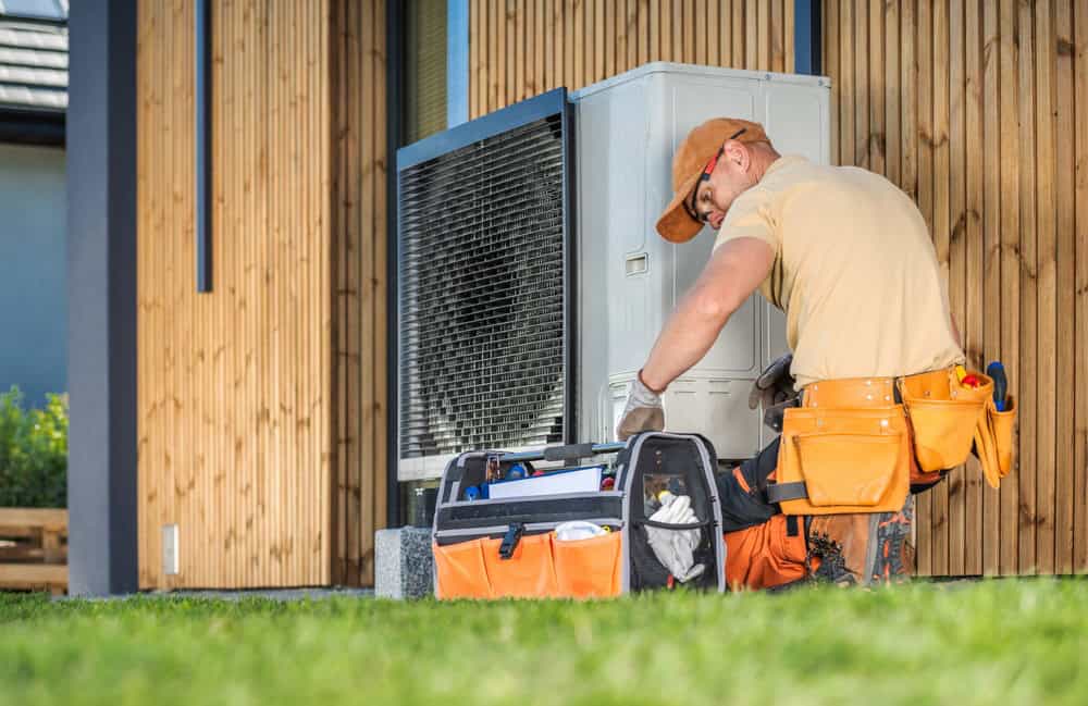 A technician wearing an orange cap works on an outdoor air conditioning unit. He kneels beside a large tool bag, surrounded by wooden paneling and grass. The scene suggests maintenance or repair work on a residential HVAC system.
