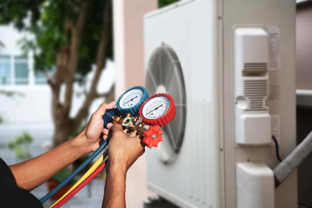 A technician uses a pressure gauge manifold to check an air conditioning unit. The image focuses on the manifold with two gauges, one blue and one red, with colorful hoses attached. The outdoor unit is in the background.