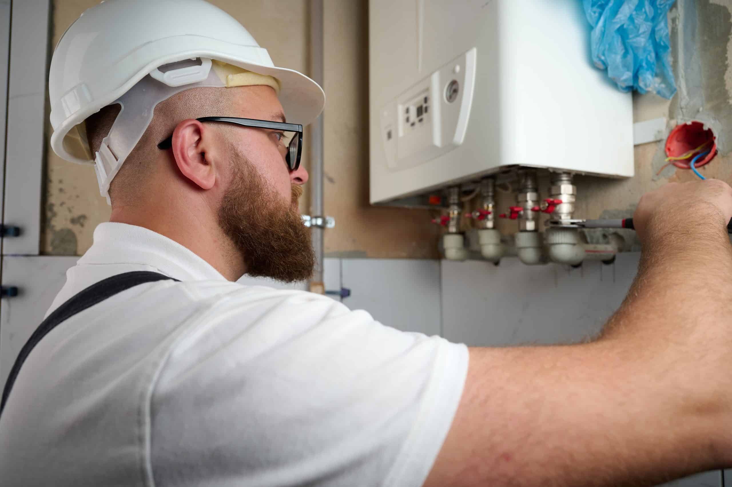 A man wearing a white hard hat and protective glasses adjusts a valve on a white wall-mounted boiler. He has a beard and is dressed in a white shirt. The setting appears to be an indoor utility or maintenance room.