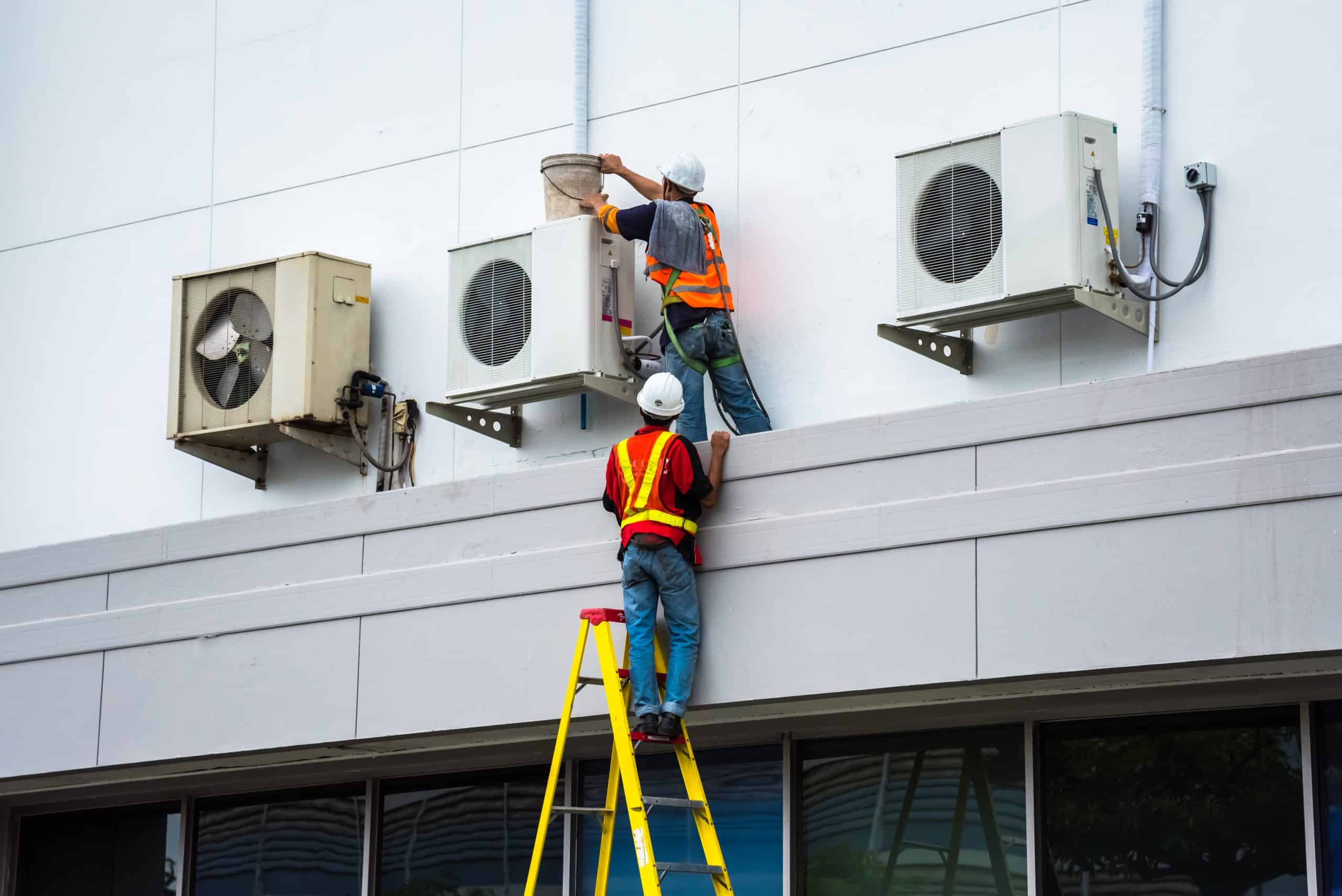 Two workers in safety gear are installing or repairing air conditioning units on an exterior wall. One stands on a ladder, while the other works directly next to the units. They both wear helmets and high-visibility vests.