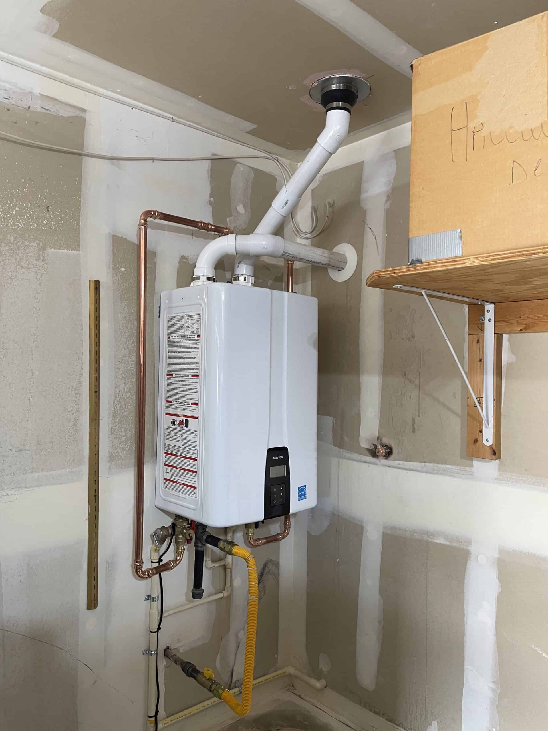 A tankless water heater is mounted on a wall with copper and PVC piping connected. A yellow gas line runs below. The surrounding walls have drywall and a shelf with a cardboard box on the right.
