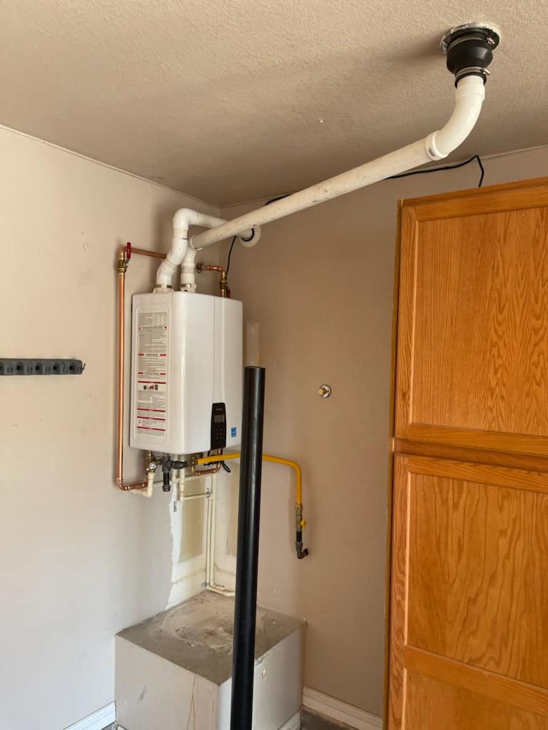 A tankless water heater is mounted on a beige wall next to a wooden cabinet. White and yellow pipes are connected to the unit, with one pipe extending towards the ceiling. A small black pole is in the foreground.