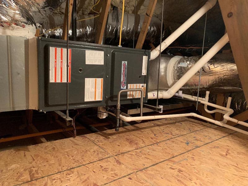A furnace and ductwork system installed in an attic with wooden flooring. Insulated pipes and multiple labels are visible on the furnace unit. The attic has sloped ceilings with visible insulation.