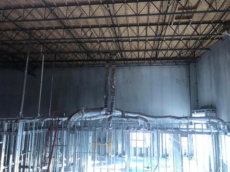 Interior of a large warehouse under construction with exposed metal beams and framing. Ductwork extends from the ceiling, and a ladder is positioned vertically. Natural light filters in from windows, illuminating the area.